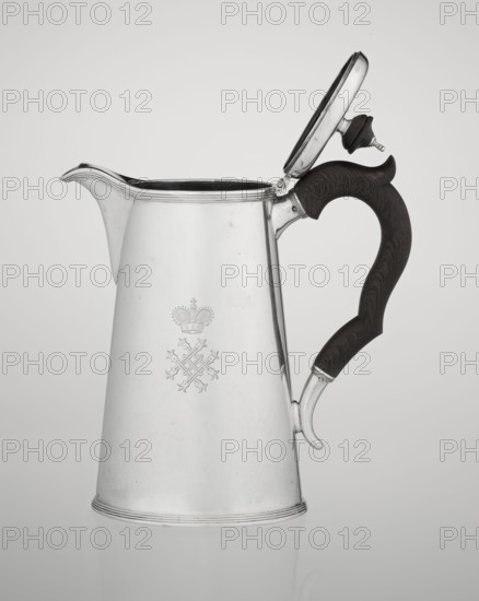 Conical coffee pot with hinged lid, slightly arched and flattened at the top, topped with a wooden handle; short spout on the axis with two sleeves for attaching a wooden handle added contemporarily. Engraved monogram tied HH under a ducal mitre; inscribed 1890/1891. Maker: Barnard, Edward & Sons Ltd London; goldsmith's firm; fl. ca. 1680– . Also associated maker/seller: Clark, Alfred Ltd London; goldsmith's firm and sellers; fl. 1898–1934. Materials: silver with wooden handle. Function: coffee ware. Place: Great Britain — England, London. Dimensions: height 17.3 cm; width 11 cm; depth 8.4 cm; weight 627 g., A metallic pitcher with a reflective silver surface and a hinged lid is shown against a light gray background, the lid open and revealing a dark interior; the pitcher has a curved spout and a dark brown wooden handle with visible grain and a carved contour, attached to the body with silver fittings, and there is an engraved decorative emblem on the pitcher’s front; the overall colo...