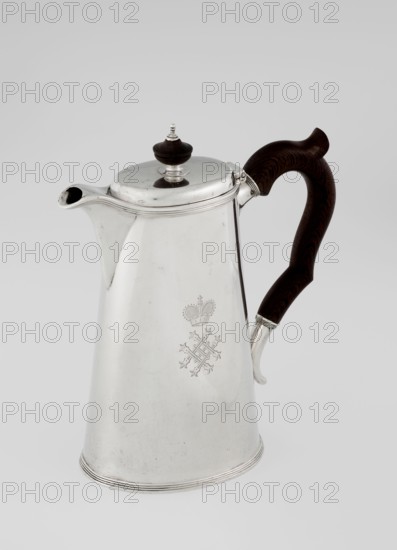 Conical coffee pot with hinged lid, slightly arched and flattened at the top, topped with a wooden handle made; spout short and axial; two sleeves on the axis for attaching a wooden handle added contemporarily. Engraved monogram tied HH under a ducal mitre; dated 1890/1891. Maker: Barnard, Edward & Sons Ltd London; goldsmiths, fl. ca. 1680– ; also associated with Clark, Alfred Ltd London; goldsmiths and sellers, fl. 1898–1934. Materials: silver with wooden handle. Function: coffee ware. Place: Great Britain — England, London. Dimensions: height 17.3 cm; width 11 cm; depth 8.4 cm; weight 627 g., A metallic pitcher with a reflective silver surface and a tapered cylindrical body sits against a plain light gray background, featuring a curved spout on one side and a dark brown wooden handle with visible grain on the other; the lid is silver with a small dark brown wooden finial topped by a metallic ornament, and the body shows a decorative engraved emblem; overall colors visible are silver,...