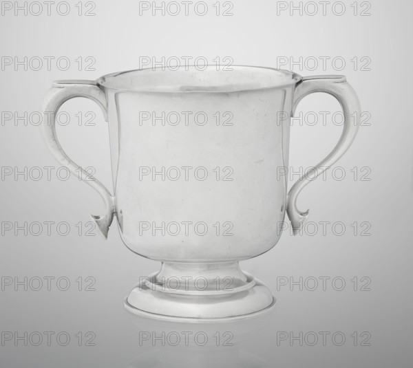 Two-eared vase, made by Hutton & Sons Ltd goldsmith and silversmith firm; fl. 1800–1930. Vase on a circular, stepped-profile foot and short stem; smooth cylindrical body with slightly upturned upper edge; two profiled volute-shaped handles on the sides. Dated 1926/1927. Great Britain — Birmingham, England. Vessel; height 15 cm, width 21 cm, depth 13.5 cm, weight 540 g., A plain metal cup with two curved handles sits centered against a smooth background; the cup has a rounded body that tapers to a stepped circular base and shows a reflective surface with slight scratches and subtle highlights, and the visible colors are shades of silver, gray, and white.