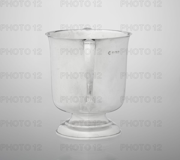 Two-eared vase, Hutton & Sons Ltd goldsmith and silversmith firm; fl. 1800–1930, vase on a circular stepped-profile foot and short stem, smooth cylindrical body with a slightly upturned upper edge, two profiled volute-shaped handles on the sides; 1926/1927; Great Britain, Birmingham, England; vessel; height 15 cm, width 21 cm, depth 13.5 cm, weight 540 g, A metallic cup-shaped vessel with a handle mounted on a rounded pedestal base, shown against a neutral light gray background; the vessel and base are reflective silver with subtle darker gray areas from reflections and surface marks, and there are a few faint irregular spots and scratches visible on the metal surface.