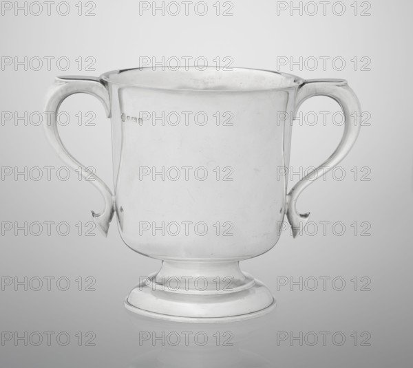 Two-eared vase by Hutton & Sons Ltd goldsmith and silversmith firm; fl. 1800–1930. Scrap-purchase provenance. Vase on a circular stepped-profile foot and short stem; smooth, cylindrical body with a slightly upturned upper edge and two profiled volute-shaped handles on the sides. Dated 1926/1927. Great Britain, Birmingham, England. Vessel; height 15 cm, width 21 cm, depth 13.5 cm, weight 540 g., A silver two-handled cup with a rounded body and a flared rim standing on a stepped circular base, showing a smooth reflective surface with light scratches and small blemishes; the handles are curving and ornate with hooked ends and are attached symmetrically at each side; the overall colors visible are shades of silver and gray with subtle highlights and shadows.