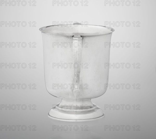 Two-eared vase, vase on a circular, stepped-profile foot and short stem, smooth, cylindrical, with a slightly upturned upper edge, two profiled, volute-shaped handles on the sides; 1926/1927; Great Britain, Birmingham, England; vessel; height 15 cm, width 21 cm, depth 13.5 cm, weight 540 g., A single metallic cup-shaped vessel with a rounded base and a short pedestal, shown from the front against a smooth light gray background; the vessel has a handle attached to its front face and a slightly flared rim, and the surface shows subtle scratches and reflections in shades of silver and gray.