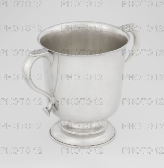 Two-eared vase, Hutton & Sons Ltd goldsmith and silversmith firm; fl. 1800–1930 maker, scrap - purchase, purchase provenance; vase on a circular stepped-profile foot and short stem, smooth cylindrical body with a slightly upturned upper edge, two profiled volute-shaped handles on the sides; 1926/1927; Great Britain, Birmingham, England; vessel; height 15 cm, width 21 cm, depth 13.5 cm, weight 540 g, A silver metal cup with a rounded body and a footed base sits against a light gray background; the cup has a smooth interior visible from above and two curved handles attached on opposite sides, each handle featuring a subtle decorative curve and a small pointed finial where it meets the body; the metal surface shows gentle reflections and a few faint scratches, and the overall colors visible are silver, gray, and the pale gray of the background.