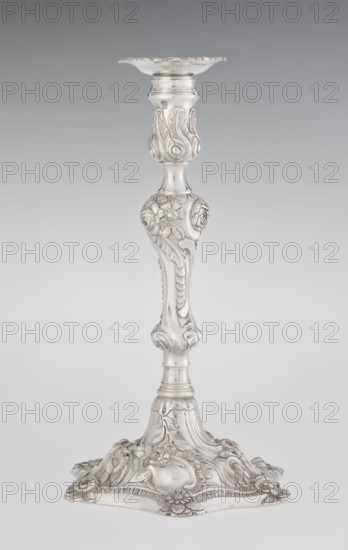 Single-candle candlestick by William Cafe fl. 1757–ca. 1802, silver, rococo/George II style, 1763/1764, London, England. Rocaille ornament; relief decoration of plant tendrils, flowers, shells, cartouches and acanthus leaves; slightly spirally twisted overall. Form: rococo on a raised domed foot with a hexagonal rim decorated with flower heads at the corners, passing into a baluster shaft and terminating in a vase-shaped socket with a disc-shaped drip pan. Height 29 cm; diameter 16.9 cm; weight 695 g., A single ornate candlestick rendered in metallic silver tones stands against a neutral grey background, featuring a scalloped dish at the top, an intricately carved shaft with floral and foliate motifs, a bulbous midsection with raised flower decorations, and a broad decorative base with flowing leaf and flower reliefs; the visible colors are silver and shades of grey.