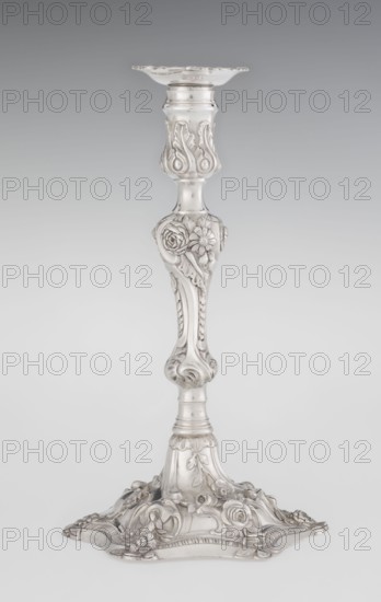 Single-candle candlestick by William Cafe fl. 1757–ca. 1802, silver, rococo George II style, 1763/1764, London, England, Great Britain. Rocaille ornament, plant tendrils, flowers, shells, cartouches, acanthus leaves; relief decoration slightly spirally twisted. Rococo on a raised domed foot with a hexagonal rim decorated with flower heads at the corners, passing into a baluster shaft and terminating in a vase-shaped socket with a disc-shaped drip pan. Function: lighting. Height 29 cm; diameter 16.9 cm; weight 695 g., A single ornate candlestick with a metallic surface in shades of silver and gray, featuring a decorative flared top, an elongated stem with sculpted floral and leaf motifs, and a wide decorative base adorned with raised flowers and scrolling details.