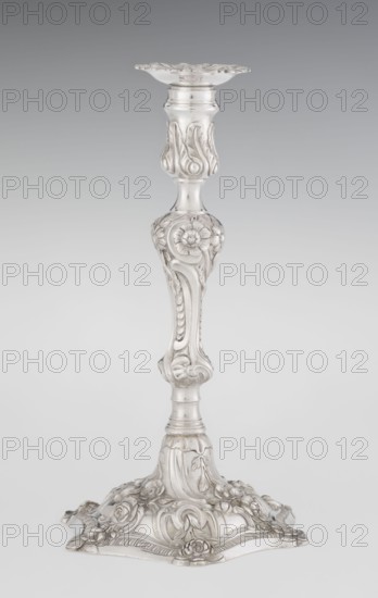 Single-candle candlestick by William Cafe fl. 1757–ca. 1802, silver, Rococo/George II style, 1763/1764, London, England. Rocaille ornament on a raised domed foot with a hexagonal rim decorated with flower heads at the corners, passing into a baluster shaft and terminating in a vase-shaped socket with a disc-shaped drip pan. Decorated with rococo motifs—rocailles, plant tendrils, flowers, cartouches, shells and acanthus leaves—with a slightly spirally twisted overall decoration. Function: lighting. Height 29 cm; diameter 16.9 cm; weight 695 g., A tall ornate candlestick rendered in silver tones stands against a plain gradient background of gray fading to a lighter gray, featuring a decorative flared top, a slender central shaft with a bulbous floral-carved midsection, and a wide sculpted base adorned with raised floral and scroll motifs; the surface shows smooth polished areas and detailed carved reliefs with visible highlights and soft shadows in shades of silver, gray, and muted white...