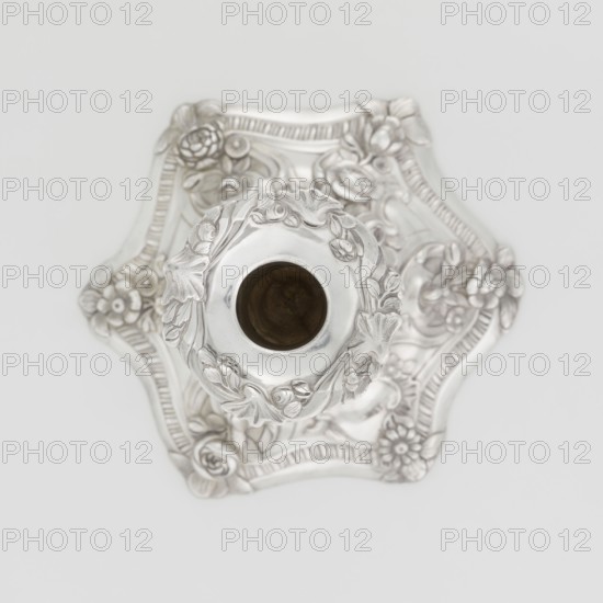 Single-candle candlestick by William Cafe fl. 1757–ca. 1802, silver, 1763–1764, London, England. Rococo George II style: raised domed foot with hexagonal rim decorated with flower heads at the corners, passing into a baluster shaft and terminating in a vase-shaped socket with a disc-shaped drip pan. Ornament includes rocailles, plant tendrils, flowers, cartouches, shells, acanthus leaves and relief decoration; overall decoration is slightly spirally twisted. Function: lighting. Height 29 cm; diameter 16.9 cm; weight 695 g., A silver ornate object viewed from above with a central circular opening surrounded by raised floral and leaf decorations, resting on a square-ish base with scalloped edges and embossed floral motifs; the visible colors are silver and a dark brownish tone inside the central opening, against a white background.