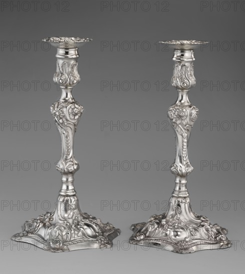Single-candle candlestick by William Cafe fl. 1757–ca. 1802, rococo; silver. Rocaille ornament; plant ornaments, flowers, shells, cartouches, acanthus leaves; relief decoration. Rococo on a raised, domed foot with a hexagonal rim, decorated with flower heads at the corners, passing into a baluster shaft, terminating in a vase-shaped socket with a disc-shaped drip pan. Decoration slightly spirally twisted in George II / Georgian style. Dated 1763/1764. Place of origin: London, England, Great Britain. Function: lighting. Height 29 cm; diameter 16.9 cm; weight 695 g., A pair of ornate metallic candlesticks photographed against a plain gray background, each with a decorative flared top, a slender stem adorned with sculpted floral and foliate motifs, and a wide ornate base with raised floral and scrollwork details; the metal has a reflective silver tone with highlights and soft shadows showing its three-dimensional relief, and the background and surface tones are various shades of gray.