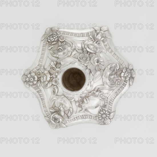 Single-candle candlestick by William Cafe fl. 1757–ca. 1802, silver, 1763/1764. Rococo George II style: rocaille ornament, plant tendrils, flowers, shells, cartouches, acanthus leaves; relief decoration slightly spirally twisted. Rococo candlestick on a raised domed foot with hexagonal rim decorated with flower heads at the corners, passing into a baluster shaft and terminating in a vase-shaped socket with a disc-shaped drip pan. Single-candle lighting object. London, England. Height 29 cm; diameter 16.9 cm; weight 695 g., A decorative metallic object with an ornate floral relief, shown from above; the piece is primarily silver with a darker brownish interior visible at its center, and it features a scalloped hexagonal shape, raised flowers and leaves, curved lines and ribbed borders, all rendered in silver tones against a light gray background.