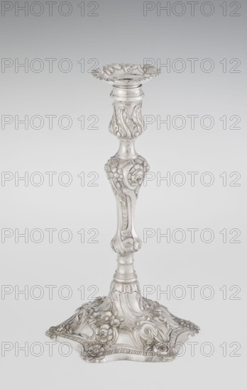 Single-candle candlestick by William Cafe fl. 1757–ca. 1802. Rococo George II style, rocaille ornament; decorated with plant tendrils, flowers, cartouches, shells, acanthus leaves and rocaille motifs. Rococo on a raised, domed foot with a hexagonal rim, flower heads at the corners, passing into a baluster shaft, terminating in a vase-shaped socket with a disc-shaped drip pan; entire decoration slightly spirally twisted. Dated 1763/1764. Great Britain — England, London. Function: lighting. Height 29 cm; diameter 16.9 cm; weight 695 g., A tall ornate metal candlestick with a shiny silver surface, featuring a scalloped top dish and a sculpted central shaft decorated with floral motifs and flowing leaf designs, a curved midsection with raised floral relief and rounded details, and a wide decorative base embellished with embossed flowers and swirling patterns; the visible colors are silver and subtle variations of gray.