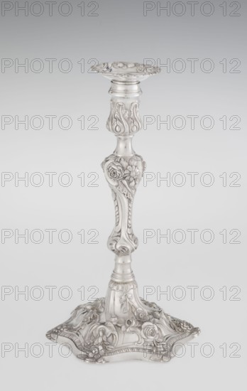 Single-candle candlestick by William Cafe fl. 1757–ca.1802, goldsmith. Rococo/Georgian George II style, 1763–1764, London, England, Great Britain. Raised domed foot with hexagonal rim decorated with flower heads at the corners, passing into a baluster shaft and terminating in a vase-shaped socket with a disc-shaped drippan. Decorated in rocaille motifs—plant tendrils, flowers, cartouches, shells, acanthus leaves—in slight spiral twist. Materials/technique: silver goldsmith work. Function: lighting. Height 29 cm; diameter 16.9 cm; weight 695 g., A tall ornate metallic candlestick stands upright against a pale gray background, showing a polished silver surface with decorative reliefs. The top has a shallow scalloped dish with floral and scroll motifs, leading to a slender stem that features raised acanthus-like leaves and rounded fluted sections. The midsection bulges into a heart-shaped knop decorated with sculpted roses and other floral forms, then tapers into a column with carved ridg...