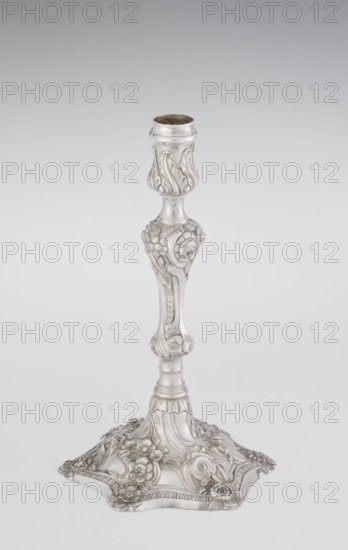 Single-candle candlestick by William Cafe fl. 1757–ca. 1802. Rococo/Georgian George II style, 1763/1764. Silver, single-candle candlestick on a raised, domed foot with a hexagonal rim, decorated with flower heads at the corners, passing into a baluster shaft and terminating in a vase-shaped socket with a disc-shaped drip pan. Ornament includes rocailles, plant tendrils, flowers, cartouches, shells, acanthus leaves and spiral-twisted relief decoration. Height 29 cm; diameter 16.9 cm; weight 695 g. Origin: London, England., A single ornate metal candlestick stands against a pale gray background, displaying a silvery finish with subtle darker tarnished areas; the upper socket has a flared rim and a bulbous section with raised leafy motifs, the slender stem features sculpted floral and scroll details and a few rounded nodes, and the broad base is shaped with curved edges and adorned with several raised flowers, leaves, and flowing decorative patterns in matching silver tones.