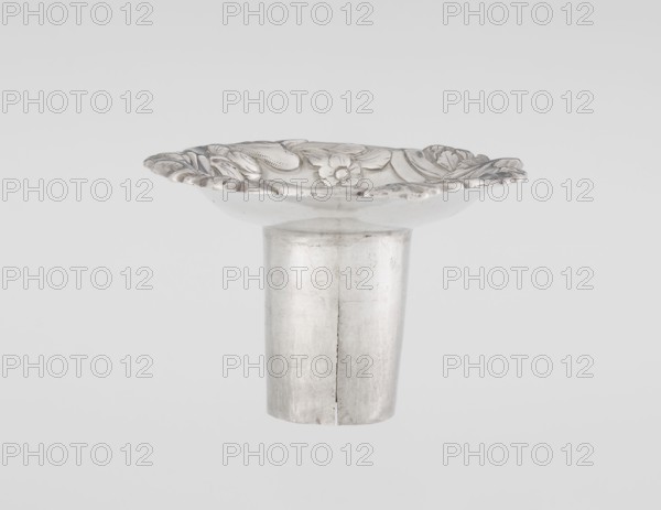 Single-candle candlestick by William Cafe fl. 1757–ca. 1802. Rococo George II style single-candle candlestick on a raised domed foot with a hexagonal rim, decorated with flower heads at the corners, passing into a baluster shaft and terminating in a vase-shaped socket with a disc-shaped drip pan. Decorated with rocailles, plant tendrils, flowers, cartouches, shells and acanthus leaves; overall decoration is slightly spirally twisted. Materials/technique: scrap. Date: 1763/1764. Place of origin: London, England, Great Britain. Function: lighting. Height 29 cm; diameter 16.9 cm; weight 695 g., A metallic object with a central cylindrical base and a shallow, flared top is centered against a light gray background; the top rim shows an ornate raised floral and leaf pattern with curved, scalloped edges, and the surface of the object appears reflective with visible scratches and subtle variations in shading; colors visible include silver, light gray, and subtle darker gray tones.