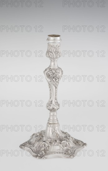 Single-candle candlestick by William Cafe fl. 1757–ca. 1802, silver, 1763/1764. Rococo George II style: rocaille ornament, plant tendrils, flowers, shells, cartouches and acanthus leaves in slight spiral twist. On a raised domed foot with hexagonal rim decorated with flower heads at the corners, passing into a baluster shaft and terminating in a vase-shaped socket with a disc-shaped drip pan. Height 29 cm; diameter 16.9 cm; weight 695 g. Origin: London, England., A single ornate metallic candlestick stands upright against a smooth pale gray background, featuring a reflective silver surface with sculpted floral and leaf motifs along its shaft and broad decorative base; the design includes rounded and curved forms, embossed roses and petals, and flowing scrollwork, all rendered in shades of silver and gray.