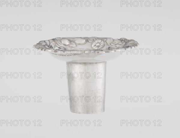 Single-candle candlestick by William Cafe fl. 1757–ca. 1802, rococo/Georgian George II style, 1763/1764, London, England. Silver single-candle candlestick on a raised domed foot with a hexagonal rim, decorated with flower heads at the corners, passing into a baluster shaft and terminating in a vase-shaped socket with a disc-shaped drip pan. Ornament includes rocailles, plant tendrils, flowers, cartouches, shells, acanthus leaves and slight spiral twisting of the overall decoration. Height 29 cm; diameter 16.9 cm; weight 695 g., A metallic object with a cylindrical base supporting a shallow, scalloped dish-like top decorated with raised floral and leaf motifs; the surface shows smooth and slightly scratched textures. Colors visible are silver and shades of gray against a pale gray background.