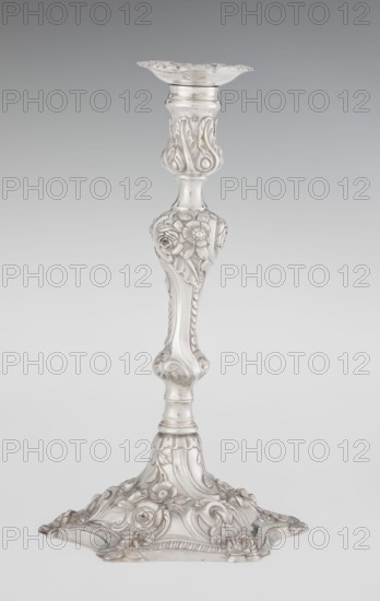 Single-candle candlestick by William fl. 1757–ca. 1802, rococo style, 1763/1764, England, London, Great Britain. Rococo single-candle candlestick on a raised, domed foot with a hexagonal rim, decorated with flower heads at the corners, passing into a baluster shaft and terminating in a vase-shaped socket with a disc-shaped profit. Decorated with rococo motifs in the George II style — rocailles, plant tendrils, flowers, cartouches, shells and acanthus leaves; entire decoration slightly spirally twisted. Materials/technique: scrap goldsmith. Function: lighting. Height 29 cm; diameter 16.9 cm; weight 717 g., A single ornate candlestick displayed against a neutral background, made of a shiny silver material with a reflective surface; it has a flared top and a tapered shaft featuring sculpted floral and leafy reliefs around the midsection and upper stem, including rose-like blossoms and petal motifs; the lower stem curves into a rounded knop with carved scrollwork and vegetal details, leadi...