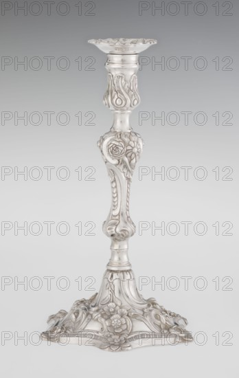 Single-candle candlestick, William fl. 1757–ca. 1802 goldsmith, rococo, scrap - purchase, plant ornaments, shells ornament, rocaille ornament, cartouches, flowers ornament, Georgian, George II style. Rococo single-candle candlestick on a raised, domed foot with a hexagonal rim, decorated with flower heads at the corners, passing into a baluster shaft, terminating in a vase-shaped socket with a disc-shaped drip pan. Decorated with rocailles, plant tendrils, flowers, cartouches, shells and acanthus leaves; entire decoration slightly spirally twisted. 1763/1764, London, England. Function: lighting. Height 29 cm; diameter 16.9 cm; weight 717 g., A tall ornate candlestick rendered in metallic silver with a glossy finish, showing a flared top with scalloped edges, a narrow neck with carved leaflike motifs, a central section with raised floral decorations and rounded contours, a lower shaft with scrolling and ribbed details, and a broad decorative base adorned with embossed flowers and flowin...