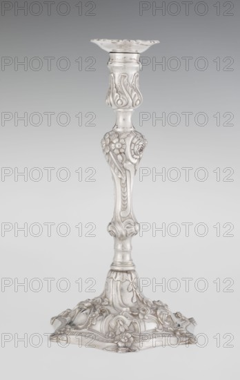 Single-candle candlestick by William Cafe fl. 1757–ca. 1802, rococo style, 1763/1764, London, England. Silver scrap construction, rococo on a raised, domed foot with a hexagonal rim decorated with flower heads at the corners, passing into a baluster shaft, terminating in a vase-shaped socket with a disc-shaped drip pan. Decorated with rococo motifs in the George II manner—rocailles, plant tendrils, flowers, cartouches, shells and acanthus leaves; the entire decoration is slightly spirally twisted. Function: lighting. Height 29 cm; diameter 16.9 cm; weight 717 g., A tall ornate candlestick made of a reflective silvery material stands against a smooth gray background, with a flared scalloped top, an elaborately carved central shaft featuring floral and leaf motifs, and a wide decorative base adorned with raised flowers, scrollwork, and curved panels; the visible colors are silver and shades of gray.