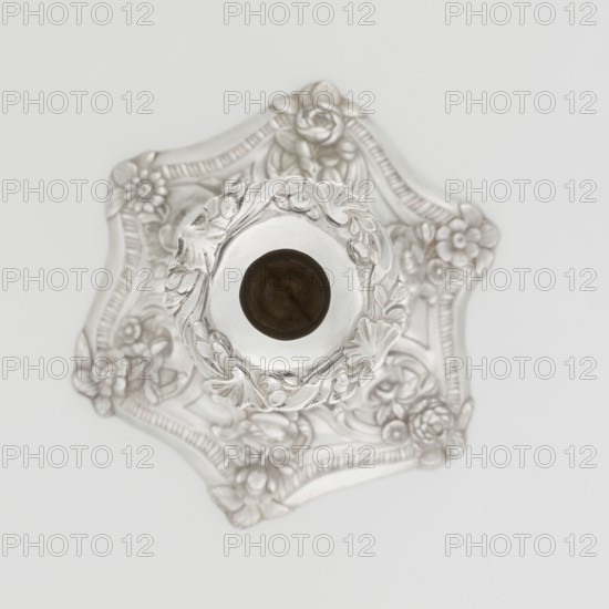 Single-candle candlestick, William fl. 1757–ca. 1802 goldsmith, rococo, scrap - purchase, plant ornaments, shells ornament, rocaille ornament, cartouches, flowers ornament, Georgian, George II style. Rococo single-candle candlestick on a raised domed foot with a hexagonal rim, decorated with flower heads at the corners, passing into a baluster shaft, terminating in a vase-shaped socket with a disc-shaped drip pan. Decorated with rocailles, plant tendrils, flowers, cartouches, shells and acanthus leaves; the entire decoration is slightly spirally twisted. 1763/1764, London, England. Function: lighting. Height 29 cm; diameter 16.9 cm; weight 717 g., The image shows a metallic decorative object with ornate floral and leaf patterns, viewed from above; the object has a central circular opening surrounded by raised floral relief and sits on a scalloped base with embossed flowers and curved lines; the visible colors are silver, white, and a darker brownish tone inside the central opening.