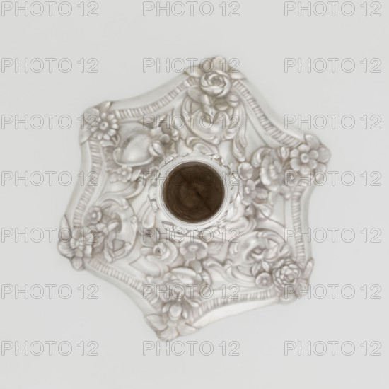 Single-candle candlestick by William Cafe fl. 1757–ca. 1802, rococo, Georgian/George II style, 1763/1764, London, England. Silver candlestick on a raised, domed foot with a hexagonal rim decorated with flower heads at the corners, passing into a baluster shaft and terminating in a vase-shaped socket with a disc-shaped drip pan. Decorated with rococo motifs — rocailles, plant tendrils, flowers, cartouches, shells and acanthus leaves — with a slight spiral twist to the overall decoration. Function: lighting. Height 29 cm; diameter 16.9 cm; weight 717 g., A decorative object viewed from above showing a sculpted base with an ornate floral and scrollwork pattern surrounding a central cylindrical opening; the surface appears metallic with shades of silver and pale gray, and the interior of the opening is darker brown.