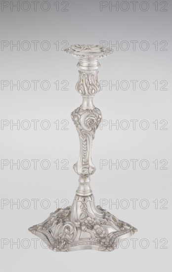 Single-candle candlestick, William Cafe fl. 1757–ca. 1802, goldsmith; rococo style, Georgian/George II influence; 1763–1764, London, England. Silver scrap-purchase technique. Rococo on a raised domed foot with hexagonal rim, decorated with flower heads at the corners, passing into a baluster shaft and terminating in a vase-shaped socket with a disc-shaped drip pan. Decorated with rocailles, plant tendrils, flowers, cartouches, shells and acanthus leaves; entire decoration slightly spirally twisted. Function: lighting. Height 29 cm; diameter 16.9 cm; weight 717 g., A single ornate candlestick stands against a light gray background, rendered in a silvery metallic tone. The piece has a flared, scalloped top with flowing carved edges, a slender central column with bulbous and contoured sections, and decorative floral and foliate reliefs along the shaft. The base is wide and irregularly shaped with raised floral motifs and curved, embossed patterns. The visible colors are silver and subtle ...
