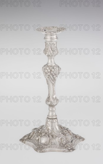 Single-candle candlestick, maker: William Cafe fl. 1757–ca. 1802, rococo style, 1763/1764, England — London, Great Britain. Single-candle candlestick on a raised, domed foot with a hexagonal rim, decorated with flower heads at the corners, passing into a baluster shaft and terminating in a vase-shaped socket with a disc-shaped profilm profit. Candlestick decorated with rococo motifs in the George II style — rocailles, plant tendrils, flowers, cartouches, shells and acanthus leaves; the entire decoration is slightly spirally twisted. Materials/technique: scrap goldsmith. Function: lighting. Dimensions: height 29 cm, diameter 16.9 cm, weight 717 g., A silver-colored ornate candlestick with a decorative round top and a flared, scalloped rim, a tapered shaft featuring raised floral and leaf motifs, a bulbous midsection with embossed flowers and scrolling details, and a wide decorative base with embossed flowers, leaves, and curved ornamental patterns, all shown against a pale gray to white...