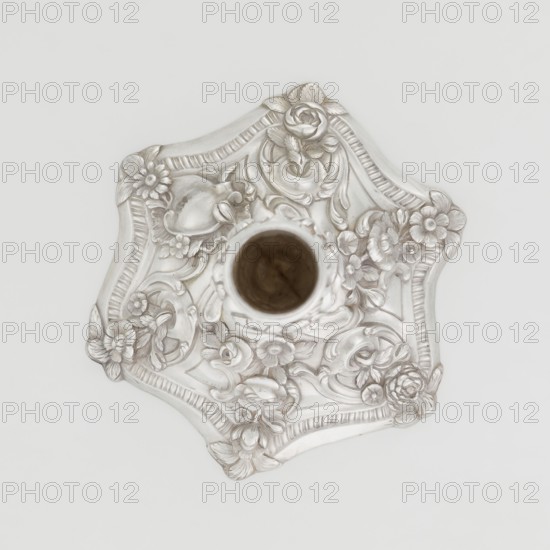 Single-candle candlestick by William Café fl. 1757–ca. 1802, rococo, 1763–1764, England, London. Rococo single-candle candlestick on a raised domed foot with a hexagonal rim, decorated with flower heads at the corners, passing into a baluster shaft and terminating in a vase-shaped socket with a disc-shaped drip pan. Decorated with rococo motifs in the George II style — rocailles, plant tendrils, flowers, cartouches, shells and acanthus leaves — with the entire decoration slightly spirally twisted. Materials/technique: scrap. Function: lighting. Dimensions: height 29 cm, diameter 16.9 cm, weight 717 g., A decorative metallic object with an ornate floral relief seen from above, featuring a central cylindrical opening and a scalloped edge; the surface shows raised sculpted flowers, leaves, and scrollwork in a silvery finish with subtle darker shadows and a small area of brown visible inside the central opening, all set against a pale off-white background.
