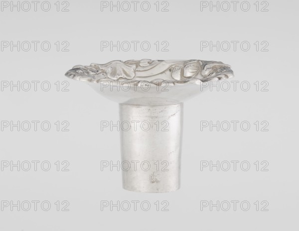 Single-candle candlestick, maker William fl. 1757–ca. 1802, rococo style; scrap — purchase; decorated with plant ornaments, shells, rocailles, cartouches, flowers and acanthus leaves. Rococo piece on a raised domed foot with a hexagonal rim, flower heads at the corners, passing into a baluster shaft and terminating in a vase-shaped socket with a disc-shaped drip pan. Decoration slightly spirally twisted. Date: 1763/1764. Place of origin: London, England, Great Britain. Function: lighting. Height: 29 cm; diameter: 16.9 cm; weight: 717 g., A single metallic object with a cylindrical stem supporting a shallow, round dish-like top that has a raised, ornate rim with curved, organic-looking embossed details; the surface appears smooth and reflective with some visible scuffs and slight texture variations. Colors visible include silver tones and subtle gray variations against a pale gray background.