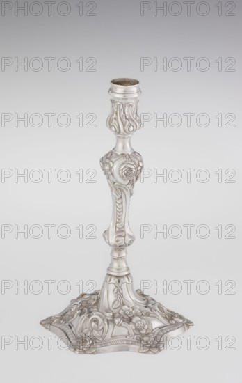 Single-candle candlestick, William Cafe fl. 1757–ca.1802, rococo George II style. Silver scrap—single-candle candlestick on a raised, domed foot with a hexagonal rim decorated with flower heads at the corners, passing into a baluster shaft and terminating in a vase-shaped socket with a disc-shaped drip pan. Decorated with rococo motifs: rocailles, plant tendrils, flowers, cartouches, shells and acanthus leaves; overall decoration slightly spirally twisted. Dated 1763/1764. London, England. Lighting. Height 29 cm; diameter 16.9 cm; weight 717 g., A single ornate candlestick stands against a plain light gray background; the candlestick is metallic silver in color with a polished sheen and features intricate carved floral and leaf motifs along its shaft and base, including rounded shapes, scrolling patterns, and raised rose-like flowers; the base spreads outward into a decorative, scalloped form with more floral relief and curved accents, and the top has a cylindrical holder with a slight...