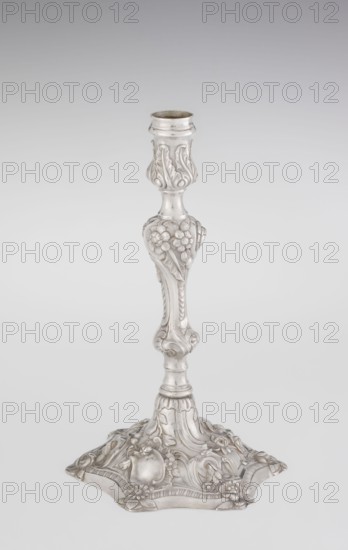 Single-candle candlestick, William Cafe fl. 1757–ca. 1802, rococo/George II style, 1763–1764, London, England. Silver candlestick on a raised, domed foot with a hexagonal rim, decorated with flower heads at the corners, passing into a baluster shaft and terminating in a vase-shaped socket with a disc-shaped drip pan. Ornamented with rococo motifs — rocailles, plant tendrils, flowers, cartouches, shells and acanthus leaves — the entire decoration is slightly spirally twisted. Function: lighting. Height 29 cm; diameter 16.9 cm; weight 717 g., A single ornate metallic candlestick stands against a pale gradient background, showing a silvery surface with decorative raised floral and leaf motifs along its shaft and base; the stem tapers and bulges in sections with carved flower clusters and flowing scrollwork, and the broad foot is shaped with curved edges and embellished with more floral reliefs and rounded forms, all rendered in shades of silver and light gray.
