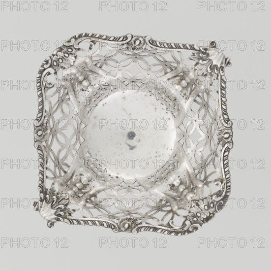 Openwork basket by Comyns & Sons London; goldsmiths' firm, fl. 1859–1953. Openwork basket on a circular foot, four-sided openwork, decorated with a motif of four-leaf rosettes and smooth stylised leaves and ribbons in the corners; irregularly cut edge enclosed in a godron half-cylinder and decorated with acanthus leaves. Materials/ornament: openwork, acanthus leaves, godron ornaments. Date: 1904–1905. Place: London, England, Great Britain. Function: vessel. Dimensions: height 4.5 cm; width 15 cm; depth 15 cm; weight 155 g., A square, ornate metal dish with an open lattice pattern and decorative scrolling edges in silver and gray tones, featuring a central shallow well with some dark speckles and a few small darker marks, rounded corner embellishments with raised bead-like forms and shell-like motifs, and an overall polished yet slightly tarnished appearance against a light gray background.