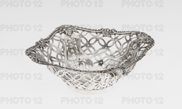 Openwork basket by Comyns & Sons goldsmiths’ firm, active 1859–1953. Four-sided openwork basket on a circular foot, decorated with four-leaf rosette motifs, smooth stylized leaves and ribbons in the corners; irregularly cut edge enclosed in a godron half-cylinder and decorated with acanthus leaves. Materials/technique: openwork, godron ornaments. Date: 1904–1905. Place: London, England, Great Britain. Function: vessel. Dimensions: height 4.5 cm; width 15 cm; depth 15 cm; weight 155 g., A decorative metallic bowl with an openwork floral and geometric pattern, showing ornate raised edges with scrolling leaf and rope-like details; the bowl has a reflective silver surface with areas of darker tarnish and subtle shine, and the background is a smooth pale gray to white gradient.