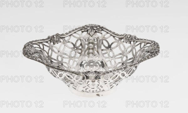Openwork basket by Comyns & Sons London; goldsmiths’ firm, fl. 1859–1953. Basket on a circular foot, four-sided openwork, decorated with a motif of four-leaf rosettes and smooth stylised leaves and ribbons in the corners; irregularly cut edge enclosed in a godron half-cylinder and decorated with acanthus leaves. Materials/technique: openwork metal with godron and acanthus ornament. Date: 1904/1905. Place: London, England, Great Britain. Classification: vessel. Dimensions: height 4.5 cm; width 15 cm; depth 15 cm; weight 155 g., A decorative metal basket with an openwork lattice pattern and ornate scrolling edges, shown against a plain light background. The basket has a shallow bowl shape with curved handles integrated into the rim and a raised central section with embossed floral and bead-like motifs. The metal surface shows polished and slightly tarnished areas. Visible colors include silver-gray tones and subtle darker gray spots.