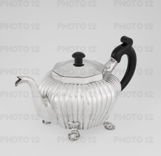 Teapot, maker: Thomas II fl. 1798–1809?, scrap silver, tea vessel. Octagonal teapot supported on four legs formed as dolphins, decorated with vertical narrow ringlets and flutes, with a spout-shaped spout, slightly arched hinged lid, wooden handle mounted in sockets not original. Inscription/date: 1808/1809. Place: London, England. Materials/measurements: silver, wooden handle; height 12 cm, width 20.5 cm, depth 14 cm, weight 595 g. Icons: dolphin motif; function: tea vessel., A silver teapot with a ribbed, rounded body sits on a surface supported by a few decorative feet; it has a curved spout and a shiny octagonal lid topped with a small black knob, and a large black handle with a curled end attached to the body. Colors visible include silver and black, with the background in light gray.