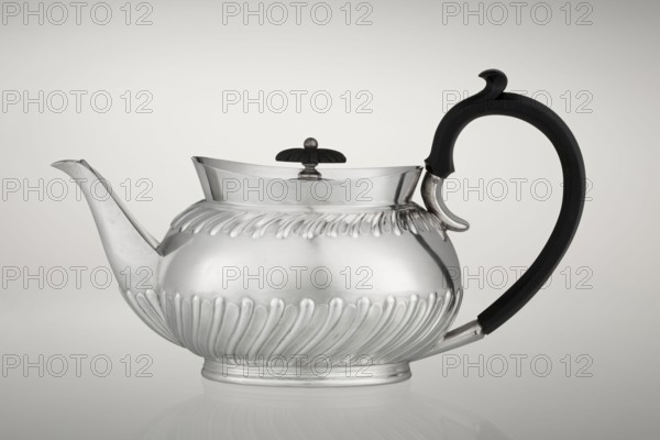 Teapot by Goldsmiths & Silversmiths Co Ltd London; makers William Gibson & John Langman goldsmiths. Egg-shaped silver teapot on an oval foot, decorated with two bands of diagonal clasp and a high upper edge, with a hinged lid topped by a handle decorated with a wooden rosette. Prominent spout on the side; non-original wooden handle set in silver sockets. Made in modern times, 1896–1897, Sheffield, England. Tea vessel tea accessory. Dimensions: height 13.5 cm, width 22.8 cm, depth 13.8 cm; weight 666 g., A polished metal teapot with a rounded, ribbed body and a short pedestal base, featuring a curved spout on one side and a large black curved handle on the other, a domed lid with a dark finial, smooth reflective silver surfaces with decorative fluting around the mid and lower sections, and visible shades of silver, black, and subtle gray tones against a pale gray background.