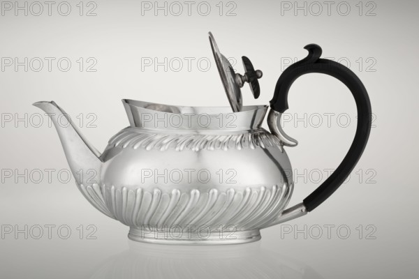 Teapot by Goldsmiths & Silversmiths Co Ltd London, makers Gibson, William & Langman, John goldsmiths. Tea vessel/teapot on an oval foot, egg-shaped, decorated with two bands of diagonal clasp, with a high upper edge, hinged lid topped with a handle decorated with a wooden rosette, prominent spout, non-original wooden handle set in sockets; made in silver, 1896/1897, Sheffield, England. Dish. Height 13.5 cm, width 22.8 cm, depth 13.8 cm, weight 666 g., A metallic teapot with a rounded, ribbed body and an open hinged lid sits on a reflective surface, showing a shiny silver finish with highlights and subtle surface imperfections; it has a long, curved spout on one side and a large, ornate handle on the other that is dark black in color and features a curved decorative top, while the lid has a small decorative knob that is also silver and dark-toned, and the background is a smooth gradient of light gray.
