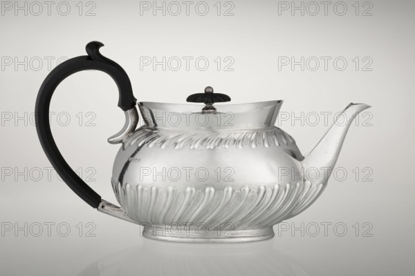 Teapot by Goldsmiths & Silversmiths Co Ltd London; makers Gibson, William & Langman, John goldsmiths. Egg-shaped tea vessel on an oval foot, decorated with two bands of diagonal clasp, high upper edge, hinged lid topped with a handle decorated with a wooden rosette; prominent spout, non-original wooden handle set in silver sockets. Made in modern times, 1896–1897, Sheffield, England. Dish. Height 13.5 cm, width 22.8 cm, depth 13.8 cm, weight 666 g., A polished metal teapot with a rounded, ribbed body and a fluted decorative band around its middle sits against a neutral pale gray background; it has a curved, elongated spout on one side and a large black handle with a hooked top on the opposite side, a small domed lid with a dark knob on top, and visible reflections and subtle surface scratches on the shiny silver metal, with colors including silver, black, and shades of gray.