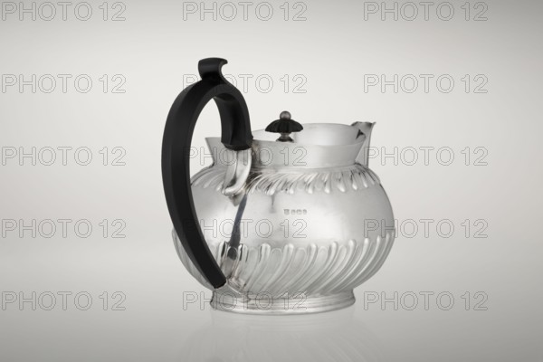 Teapot by Teapot, Goldsmiths & Silversmiths Co Ltd; makers William Gibson & John Langman goldsmiths. Egg-shaped silver teapot on an oval foot, decorated with two bands of diagonal clasp, high upper edge, hinged lid topped with a handle decorated with a wooden rosette, prominent spout, non-original wooden handle set in sockets. Made in modern times, 1896–1897, Sheffield, England. Height 13.5 cm; width 22.8 cm; depth 13.8 cm; weight 666 g., A rounded metal pitcher with a reflective silver surface and decorative fluted ridges around its lower and upper body sits against a light neutral background, featuring a curved black handle attached to one side and a scalloped spout on the opposite side, with a small dark knob on a short stem at the top; visible colors include silver, black, and shades of gray, and the pitcher rests on a slightly raised circular base with its reflection faintly visible on the surface beneath.