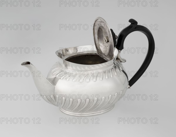 Teapot by Goldsmiths & Silversmiths Co Ltd London; makers William Gibson & John Langman goldsmiths. Egg-shaped silver teapot on an oval foot, decorated with two bands of diagonal clasp, with a high upper edge, hinged lid topped with a handle decorated with a wooden rosette, prominent spout, non-original wooden handle set in silver sockets. Made 1896–1897, Sheffield, England. Tea vessel; dish. Height 13.5 cm, width 22.8 cm, depth 13.8 cm, weight 666 g., A rounded metal teapot with a reflective silver surface and an open hinged lid sits against a pale gray background, showing a dark interior; the body features raised decorative ridges around the top and base and a short spout extending from one side; a curved black handle with a slight decorative curl at the top is attached opposite the spout.