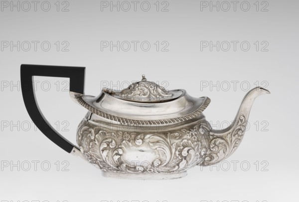 Teapot. Maker: Henry Moreton fl. 1899–1930s, factory owner, goldsmith. Neo-Rococo style; relief decoration with neo-Rococo motifs and acanthus leaves; godrons ornament. Egg-shaped teapot on an oval plinth; spout-shaped spout on the side; sleeves for attaching a wooden handle added contemporarily mounted on the axis. Upper edge bordered with a band of godrons with a wide collar above; hinged, centrally arched lid topped with a handle; oval blind reserve on the belly with acanthus rim. Date: 1909–1910. Place of origin: Chester, England, Great Britain. Function: tea vessel. Dimensions: height 13.5 cm, width 26 cm, depth 12 cm; weight 555 g., A decorative metal teapot with ornate raised floral and scrollwork patterns covering its rounded body, a curved spout with matching engraved designs, and a hinged domed lid topped with a sculpted finial; the teapot has a contrasting angular handle that appears smooth and blocky. The visible colors are silver on the metal surfaces and black on the hand...