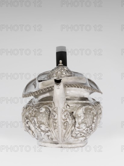 Teapot by Henry Moreton fl. 1899–1930s, factory owner and goldsmith; neo-Rococo style; silver; 1909/1910, Chester, England. Egg-shaped teapot on an oval plinth with a side spout, sleeves on the axis for a wooden handle added contemporarily, hinged centrally arched lid topped with a handle. Upper edge bordered with a band of godrons and a wide collar. Entire surface decorated in neo-Rococo relief motifs and acanthus leaves, with an oval blind reserve on the belly framed by an acanthus rim. Function: tea vessel. Height 13.5 cm; width 26 cm; depth 12 cm; weight 555 g., A silver teapot with ornate embossed floral and scrollwork decoration sits against a gray to white gradient background; the body is rounded with raised decorative panels and a sculpted vertical ridge at the front that integrates into the short spout, the hinged lid has a domed finial and a scalloped edge, and a black handle with a gray top extends upward from the back; visible colors include silver, black, gray, and white.