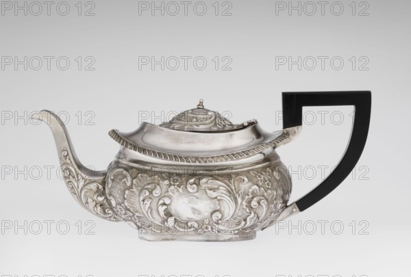 Teapot, Henry Moreton fl. 1899–1930s, factory owner; neo-Rococo style; silver scrap - purchase provenance noted; decorated with acanthus leaves and godrons; tea vessel in relief decoration; teapot on an oval plinth, egg-shaped; side spout, sleeves for attaching a wooden handle mounted on the axis added contemporarily; upper edge bordered with a band of godrons and a wide collar; hinged lid, centrally arched, topped with a handle; overall decorated with neo-Rococo relief motifs and acanthus leaves; oval blind reserve on the belly with acanthus rim; dated 1909/1910; Chester, England; height 13.5 cm, width 26 cm, depth 12 cm, weight 555 g., A decorative teapot with an ornate metallic body and a dark angular handle is shown against a pale gray background; the teapot surface is silver with embossed floral and scroll motifs covering its sides and spout, a ridged decorative rim along the top, and a domed lid with sculpted details and a small finial, while the handle is black and angular, cont...