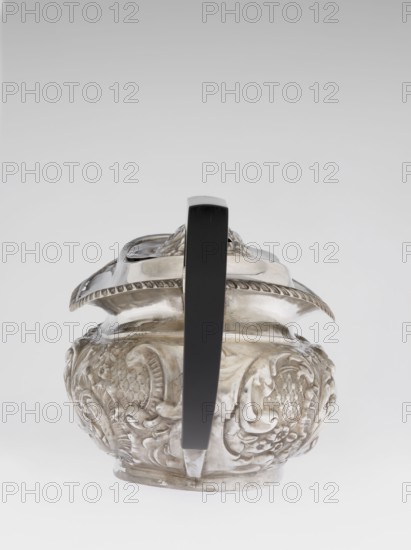 Teapot by Henry Moreton fl. 1899–1930s, factory owner and goldsmith; neo-Rococo style; silver; relief decoration with neo-Rococo motifs and acanthus leaves; teapot on an oval plinth, egg-shaped body with spout on the side; sleeves for attaching a wooden handle mounted on the axis added contemporarily; upper edge bordered with a band of godrons and a wide collar; hinged, centrally arched lid topped with a handle; oval blind reserve on the belly with acanthus rim; dated 1909/1910; Chester, England; vessel. Dimensions: height 13.5 cm, width 26 cm, depth 12 cm; weight 555 g., An ornate metal vessel with a rounded body and embossed decorative motifs sits centered in the image, viewed from the front with a tall straight handle rising from its top to bottom; the vessel surface shows intricate raised floral and scroll patterns and a scalloped rim, and the handle appears smooth and solid. The visible colors are silver on the metal surface and black on the handle, set against a pale gray to whit...