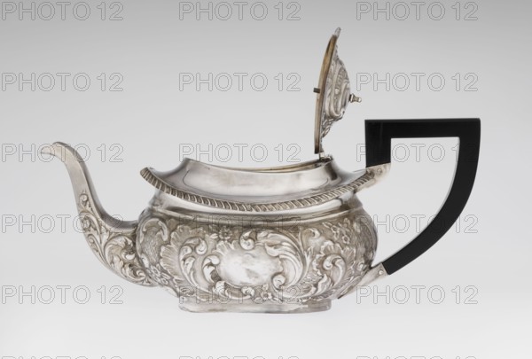 Teapot by Henry Moreton fl. 1899–1930s, maker/factory owner; neo-Rococo style; silver; relief decoration with neo-Rococo motifs and acanthus leaves; egg-shaped teapot on an oval plinth; spout-shaped spout on the side; sleeves for attaching a wooden handle added contemporarily mounted on the axis; upper edge bordered with a band of godrons, above it a wide collar; hinged lid, centrally arched, topped with a handle; oval blind reserve on the belly with acanthus rim; dated 1909/1910; Chester, England; vessel. Dimensions: height 13.5 cm, width 26 cm, depth 12 cm; weight 555 g., A silver teapot with ornate raised floral and scrollwork decoration along its rounded body, a curved spout with matching decorative motifs, a hinged lid shown open with a detailed underside, and a black angular handle attached to the body; visible colors include silver, black, and small hints of warm metal tones against a light gray background.