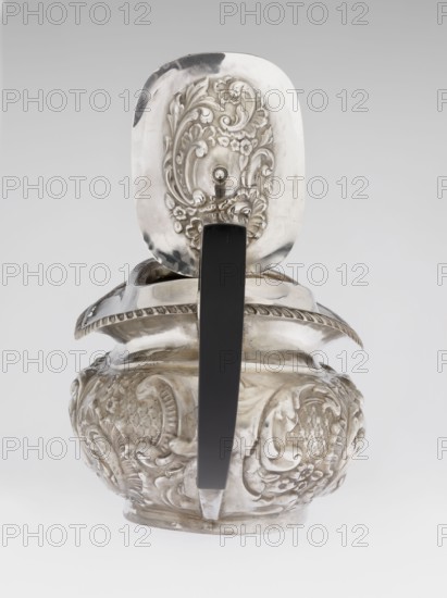 Teapot by Henry Moreton fl. 1899–1930s, maker factory owner, neo-Rococo style, silver, 1909/1910, Chester, England. Egg-shaped teapot on an oval plinth with a side spout, sleeves for attaching a wooden handle mounted on the axis handle added contemporarily. Upper edge bordered with a band of godrons and a wide collar; hinged, centrally arched lid topped with a handle. Decorated overall with neo-Rococo relief motifs and acanthus leaves; oval blind reserve on the belly with acanthus rim. Function: tea vessel. Height 13.5 cm; width 26 cm; depth 12 cm; weight 555 g., A decorative metal vessel with an ornate lid sits against a light gray background, showing intricate raised floral and scrollwork patterns on the lid and body; the vessel has a curved rim with a rope-like edge and a bulbous lower section covered in embossed motifs, and a dark vertical handle attached from the lid to the base; visible colors include silver tones on the metal, a dark black or very dark gray on the handle, and pa...