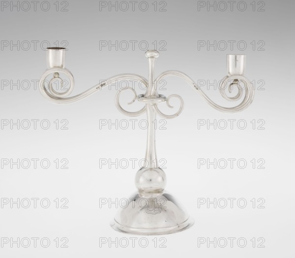 Two-candle candlestick. Maker: William Leuchars fl. 1876–1888; firm: Leuchars & Son London, goldsmiths, fl. ca. 1845–1902., silver lighting fixture, two-branched candlestick on a circular domed foot. Description: shaft rests on a ball no finial, enclosed at 3/4 height in a plate-like ring from which extend movable arms of S-shaped twisted wires, split at the ends, terminating in cylindrical candle sockets. Foot engraved with the Pilawa coat of arms with motto beneath a nine-baton count's crown; includes coats of arms and heraldic rank crowns. Date: 1881–1882. Place of origin: London, England, Great Britain. Dimensions: height 27 cm; width 32 cm; depth 11.5 cm; weight 1082 g., A silver-colored metal candelabrum with a central vertical stem rising from a rounded base, a bulbous knop near the lower stem, and two symmetrically curved arms that curl into spiral scrolls ending in cylindrical candle cups; the surface is reflective and shows highlights and subtle darker tarnish on some areas, ...