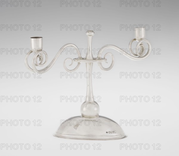 Two-candle candlestick by William Leuchars fl. 1876–1888 and Leuchars & Son London; goldsmith's firm, fl. ca. 1845–1902. Two-branched candlestick on a circular, domed foot; the shaft rests on a ball no finial, enclosed at three-quarters of its height in a plate-like ring, from which extend movable arms made of S-shaped twisted wires, split at the ends, terminating in cylindrical sockets for candles. The foot is engraved with the Pilawa coat of arms with the motto beneath a nine-baton count's crown. 1881/1882. Great Britain; England; London. Lighting. Height 27 cm; width 32 cm; depth 11.5 cm; weight 1082 g., A metallic two-armed candlestick with a rounded base and a central stem topped by a small knob sits against a neutral background; the arms curve outward into decorative spirals ending in cylindrical holders, and the object appears in shades of silver and gray with reflections and subtle highlights.