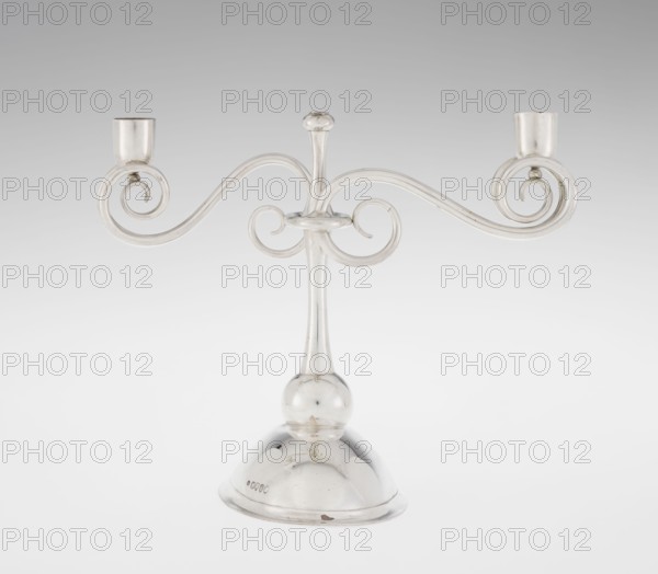 Two-candle candlestick by William Leuchars fl. 1876–1888, Leuchars & Son London, goldsmith's firm, fl. ca. 1845–1902. Two-branched candlestick on a circular domed foot. The shaft rests on a ball no finial and is enclosed at three-quarters of its height by a plate-like ring, from which extend movable arms made of S-shaped twisted wires, split at the ends and terminating in cylindrical candle sockets. The foot is engraved with the Pilawa coat of arms and motto beneath a nine-baton count's crown. Date: 1881/1882. Origin: London, England. Function: lighting. Dimensions: height 27 cm; width 32 cm; depth 11.5 cm. Weight: 1082 g., A metallic candlestick with a polished silver appearance stands on a rounded dome base and a spherical joint, rising into a slender central stem topped with a small rounded finial; two ornate horizontal arms extend from the stem in symmetrical curved scrolls ending in cup holders, creating an overall elegant, curving silhouette. The visible colors are silver and sha...