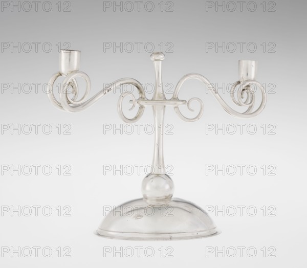 Two-candle candlestick, attributed to William Leuchars fl. 1876–1888 and the firm Leuchars & Son London; goldsmiths, fl. ca. 1845–1902. Silver two-branched candlestick on a circular domed foot. The shaft rests on a ball no finial and is enclosed at three-quarters of its height by a plate-like ring, from which extend movable arms made of S-shaped twisted wires, split at the ends and terminating in cylindrical candle sockets. The foot is engraved with the Pilawa coat of arms beneath a nine-baton count’s crown, with mottos. Date: 1881–1882. Place of origin: London, England, Great Britain. Function: lighting. Dimensions: height 27 cm; width 32 cm; depth 11.5 cm; weight 1082 g., A clear glass two-armed candlestick with a rounded dome base and a slender central stem topped by a small bulb, featuring ornate curved scroll arms that extend outward and curl into circular spirals ending in short cylindrical holders; the visible colors are clear glass with subtle highlights of white and pale gray ...
