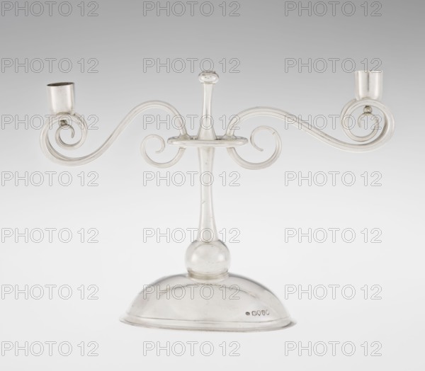 Two-candle candlestick. Maker: William Leuchars fl. 1876–1888; firm: Leuchars & Son London, goldsmiths, fl. ca. 1845–1902. Materials/technique: silver goldsmithing. Description: two-branched candlestick on a circular domed foot; shaft rests on a ball no finial, enclosed at three-quarters of its height in a plate-like ring from which extend movable arms made of S-shaped twisted wires, split at the ends, terminating in cylindrical candle sockets. Foot engraved with the Pilawa coat of arms and motto beneath a nine-baton count's crown. Date: 1881/1882. Place of origin: London, England, Great Britain. Function: lighting. Dimensions: height 27 cm; width 32 cm; depth 11.5 cm; weight 1082 g., A silver-colored metal candelabrum with a rounded base and a slender central stem topped by a small knop, from which two horizontally curving arms extend outward in symmetrical scroll shapes ending in cup-like candle holders; the surface shows subtle reflections and faint scratches, and the background is ...